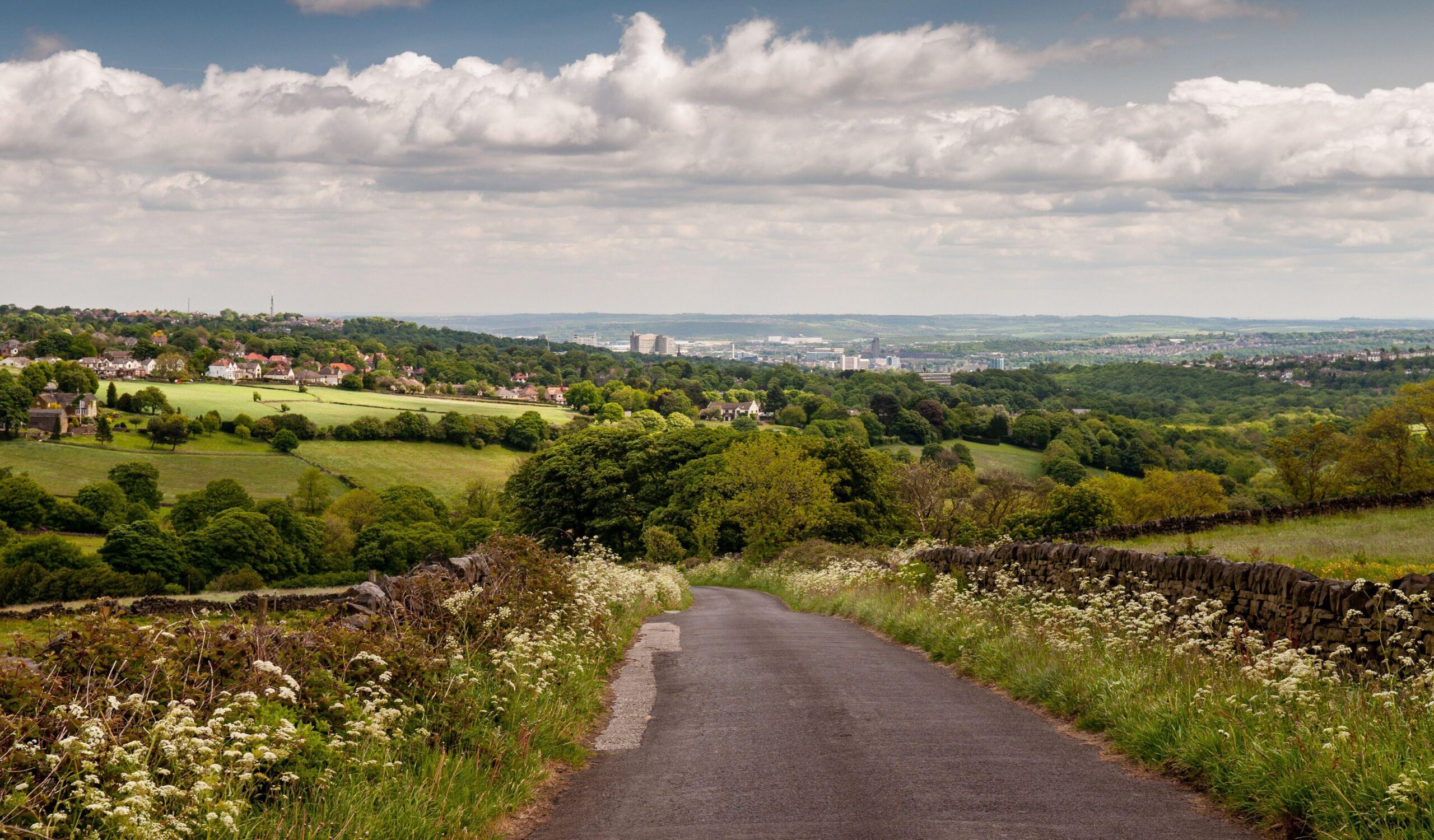 A country lane descends from the Peak District hills into the Mayfield Valley and the suburbs of Sheffield, with the city centre cityscape in the distance