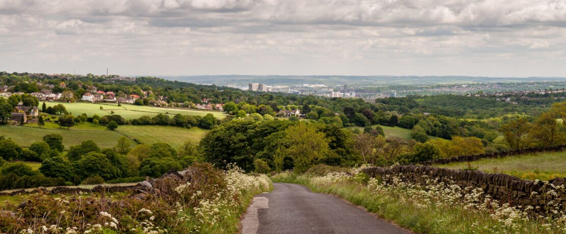 A country lane descends from the Peak District hills into the Mayfield Valley and the suburbs of Sheffield, with the city centre cityscape in the dist