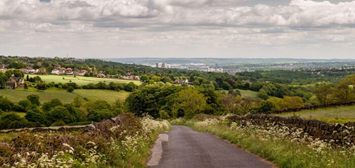 A country lane descends from the Peak District hills into the Mayfield Valley and the suburbs of Sheffield, with the city centre cityscape in the dist