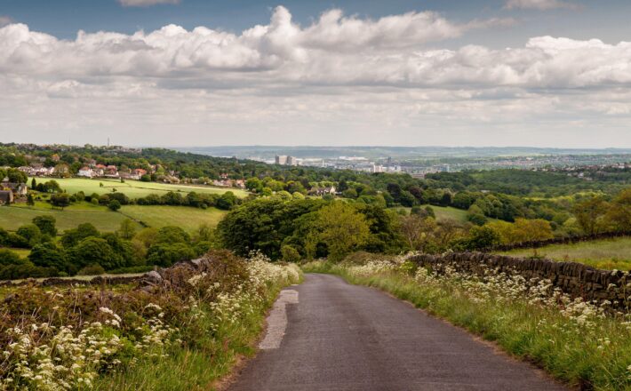 A country lane descends from the Peak District hills into the Mayfield Valley and the suburbs of Sheffield, with the city centre cityscape in the dist