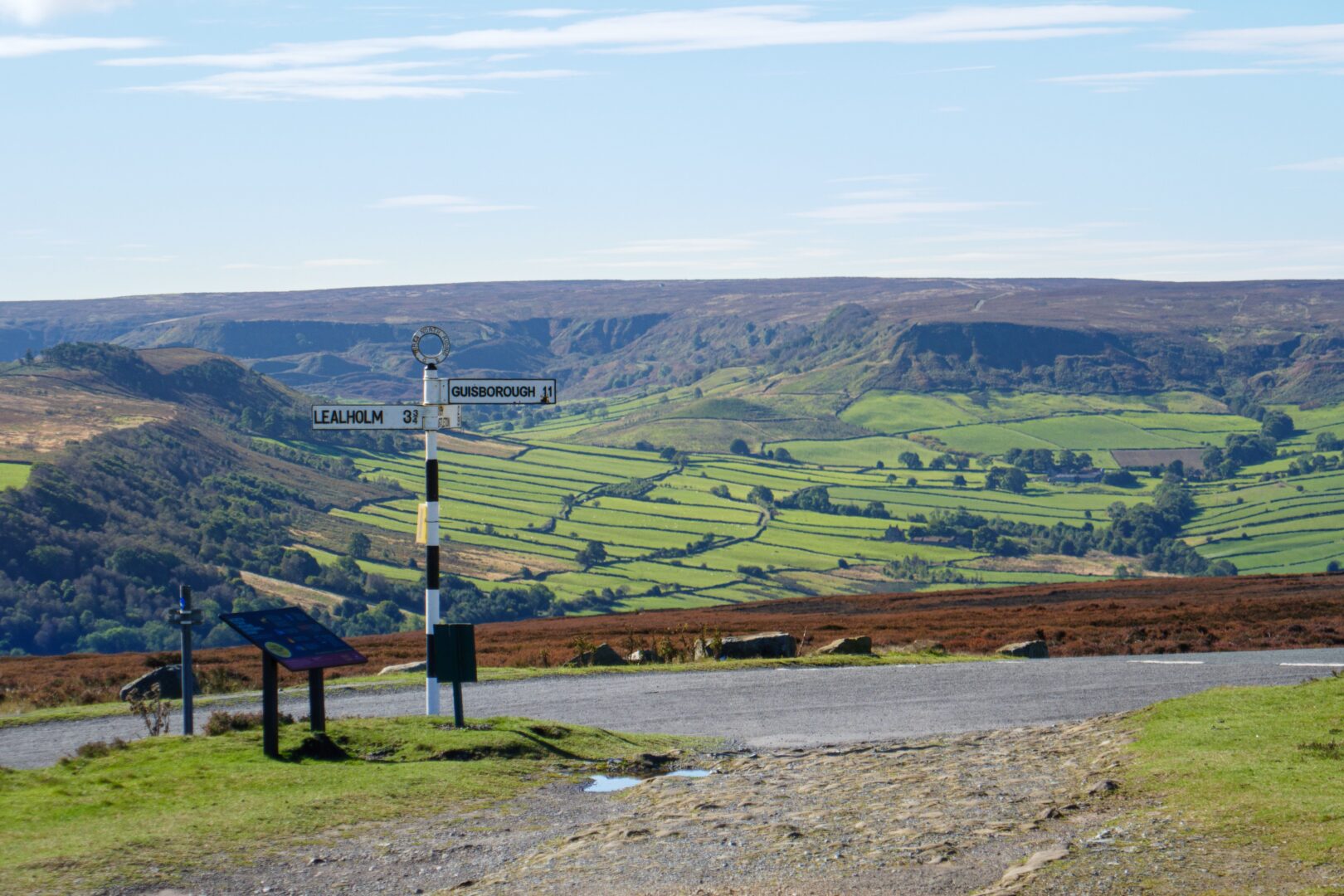 Panoramic view of North York Moors with iconic signpost, showcasing lush green landscape on a sunny day.