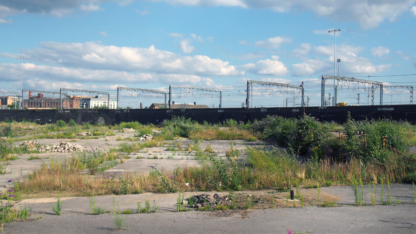 Image of a brownfield site. Image shows a large disused urban site. Covered in cracked concrete with weeds and grass growing through. In the background is a train train and some urban buildings. There is a blue sky with some scattered white clouds.
